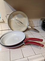 Four-piece skillet set on tiled kitchen counter: two Tasty skillets with red handles and white cooking surface, one Trisha Yearwood skillet with matching red handle and white interior, and a glass lid with metal handle propped behind.