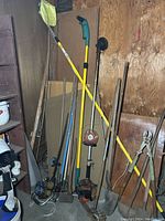 Photo showing long-handled garden tools including shovels, gas trimmers, fruit picker near corner storage unit.