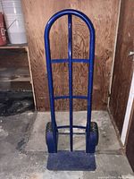 Front view of blue metal hand truck dolly showing curved handle, vertical and horizontal support bars, and base plate.