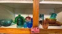 Shelf with multiple colored glassware pieces including bowls, jars, tumblers, and decorative items in various colors such as blue, green, amber, and red.