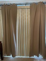 Photo showing two tan blackout drapes hanging with 8 silver rings on a metal curtain rod with vertical blinds behind.