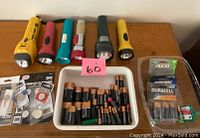 Top-down image showing six assorted flashlights lined up on a table with a tray of various new and used batteries. Batteries include AA, AAA, C, and D sizes and some button cell packages.