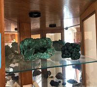 Glass cabinet displaying six geodes in green and blue-green mineral formations of various sizes and textures, some with hollow centers.