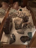 Assortment of geodes and mineral specimens arranged on white cloth inside a display cabinet. Various sizes and colors visible including white crystalline, dark brown slab, pinkish geodes, and green mineral chunks.