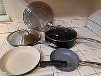 Full set of four skillets, two lids and OXO grease catcher on tiled kitchen counter.