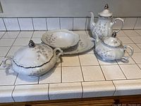 Wide view of multiple pieces on kitchen counter showing coffee pot, teapot, serving bowl, platter, and soup tureen all decorated with matching floral patterns and silver finials on lids.