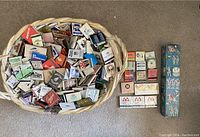 Top-down image showing woven basket filled with a large number of assorted matchboxes, scattered next to basket are additional boxed matches including brands and some decorative packaging.