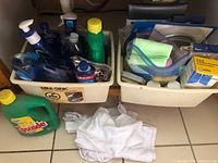 Wide view of kitchen cleaning supplies and rags under the sink, showing bottles, mat, and cloths.