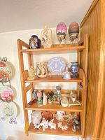 Wooden three-tier hanging display shelf with various decorative items including painted ostrich eggs, Wedgwood plate, bottle and bell, ceramic pitcher, owl figurine, and assorted figurines on shelves.