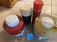 Overview of four vintage thermoses: two large Coleman jugs and two smaller personal sized. All plastic, with screw-top lids and some with handles and spouts.