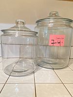 Two clear glass canisters shown side by side on a tiled surface. Both have lids with knob handles. The canister on the left is shorter and smaller than the one on the right.