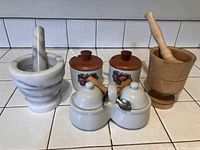 Two ceramic jam jars with lids and spoons, one grey marble mortar and pestle, and one wooden mortar and pestle arranged on a counter