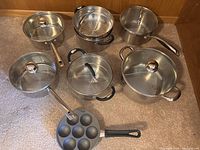 Eight-piece collection of stainless steel pans including saucepans with glass lids, steamer insert, and egg poacher pan.