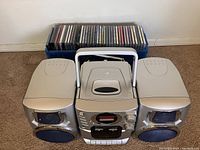 Photo showing a silver compact stereo system with a central unit featuring a CD player, radio tuner, cassette deck, and dual silver detachable speakers with black speaker mesh. A large blue container behind holds many audio CDs.