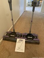 Image showing two upright Shark cordless sweepers side by side on carpet with owner's manual in front.