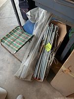 Folded clear vinyl tent or tarp with metal poles bundled inside a box beside a small plaid folding chair