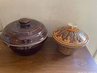 Two vintage stoneware bowls with lids, one large dark brown and one smaller decorative brown-orange, placed on a wooden surface.