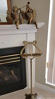 Two gold-colored resin angel statues playing wind instruments on a shelf above a fireplace, with a brass music stand positioned in front.