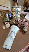 Overview of Asian themed collection including ceramic tiffin box, cloisonne vases, carved stone vases, porcelain vase, and small metal figurines on brass tray and table surface.