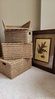 Three woven wicker storage baskets stacked and decorative framed oak leaf and acorn print leaning against the wall.