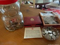 Overall lot including large bear-shaped jar filled with coins, a red coin case with papers, a wooden box with coin bags, and a small glass dish of coins on a wooden table.