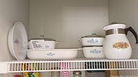 Shelf view showing white CorningWare bakeware with blue cornflower pattern, including casserole dishes with glass lids and a large oval pie plate, plus a coffee pot with fruit design