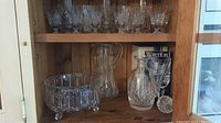 Full view of shelved crystal glassware including footed bowl, pitcher, Waterford Lismore decanter with box, and multiple glasses.