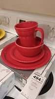 Stacked red Fiestaware dinnerware: dinner plate, side plate, bowl, with matching mug on top, displayed on stove with boxes beneath.