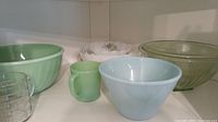 Seven assorted vintage bowls and a mug on shelf, including white scalloped Arcopal France bowl, light green Fire King Jadeite swirl mixing bowl and mug, and green ribbed uranium glass nested bowls.