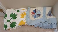 Two handmade quilts folded and leaning against a wall, one with green and yellow leaves on white, the other with pastel blue floral petal patches and scalloped edge