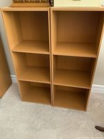 Pair of light wood-finish shelf units with three cubby compartments each, shown standing side by side on carpeted floor.