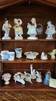 Shelf display with multiple porcelain figurines including children and birds along with small teacups and floral dish