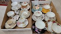 Two cardboard trays containing eleven teacup and saucer sets in various floral patterns along with creamer, sugar bowl and heart dish