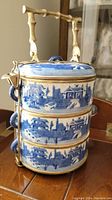 Front view of the three-tier white pottery tiffin carrier with blue Asian scenic glaze and bamboo textural brass handle.