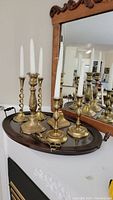 Full view of three brass candlestick holders with white candles on an oval wooden tray with glass insert, displayed in front of a mirror.