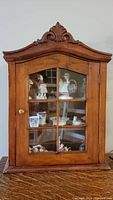 Front view of vintage wooden display cabinet showing glass front door with decorative carving on top and display items within.