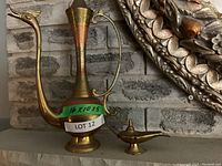 Three brass decorative items displayed against a brick background: a tall pitcher and two Aladdin-style oil lamps with engraved designs.