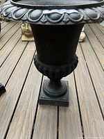 Side view of black cast iron planter urn showing fluted rounded lower portion and square pedestal base on wooden deck.