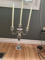 Full view of the clear crystal candelabra on wooden floor, showing three white taper candles, center taller than sides.
