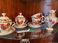 Full view of the tea set on a glass shelf showing teapots, cups, saucers, and lidded sugar bowls with red and black designs