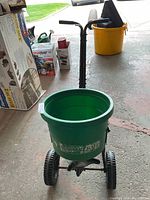 Front view of green plastic hopper lawn spreader with black handlebar and wheels.