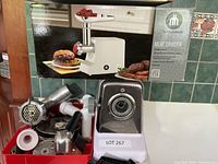 Wide shot of the meat grinder, several metal attachments, and the original product box with logo and product image.