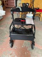 Front view of the Nexus wheelchair showing black cushioned seat and backrest, front caster wheels, and large storage bag underneath.
