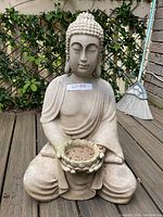 Full view of seated Buddha statue holding a planter bowl on a wooden deck with plants and broom in background.