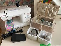 Singer Model 132 sewing machine with foot pedal and colorful fabric under needle, sewing box open behind it, and two small containers with sewing notions in front.