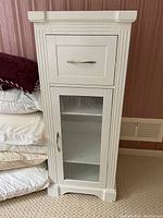 White wooden cabinet with closed drawer and closed glass door beside folded bedding.