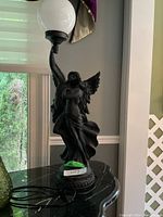 Full view of the lamp on a black marble surface showing the sculpted angel figures holding the white spherical lampshade.