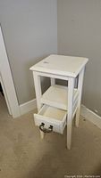 White painted wooden bedside table showing drawer open and bottom shelf, positioned near wall outlet.