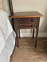 Full view of wooden bedside table with two drawers next to a bed.