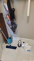 Photo showing flat floor mop with long handle, small electric iron, blue plastic watering can, and two detergent bottles and bleach bottle on floor.